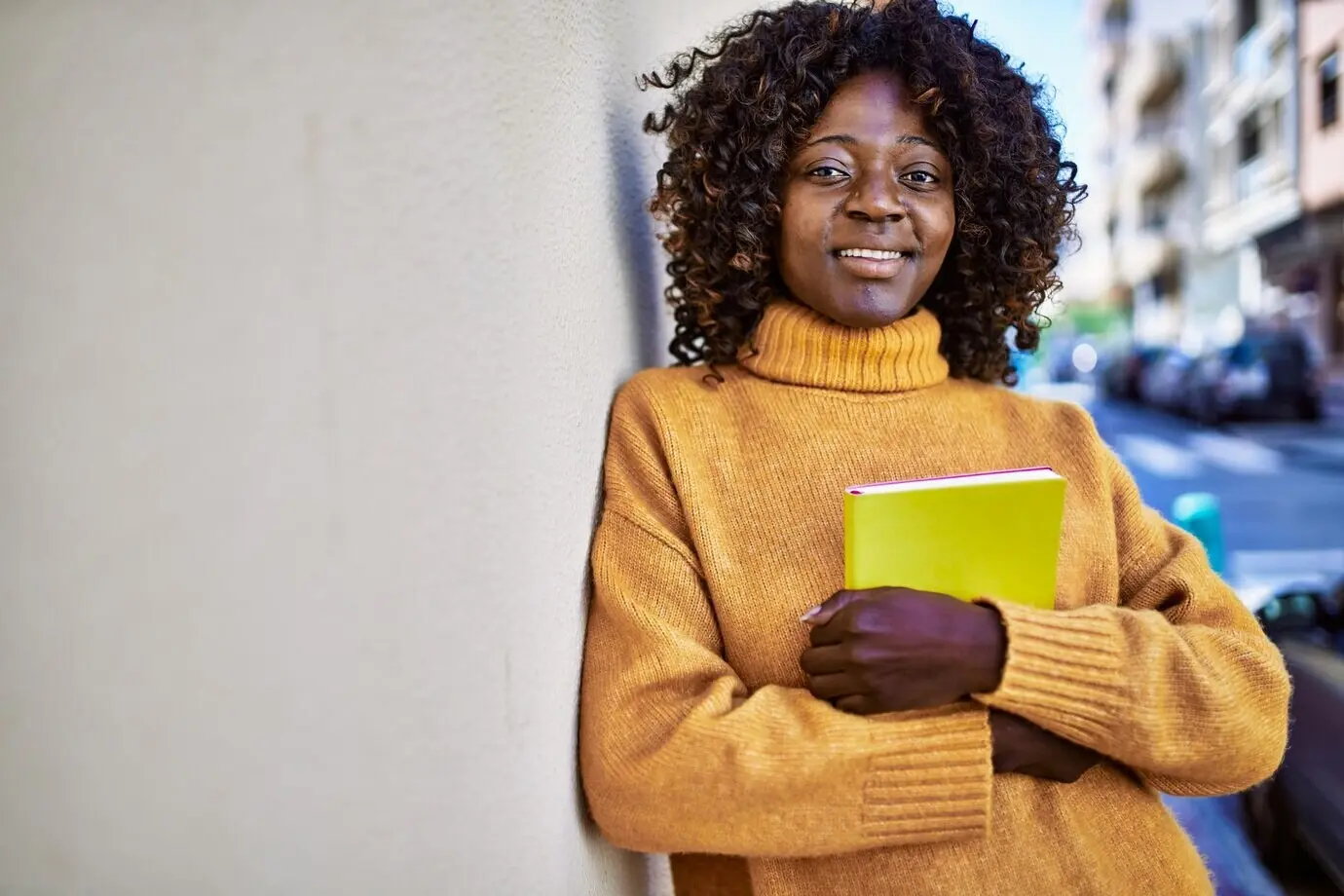 Eine afroamerikanische Frau lächelt selbstbewusst und hält auf der Straße ein Buch.